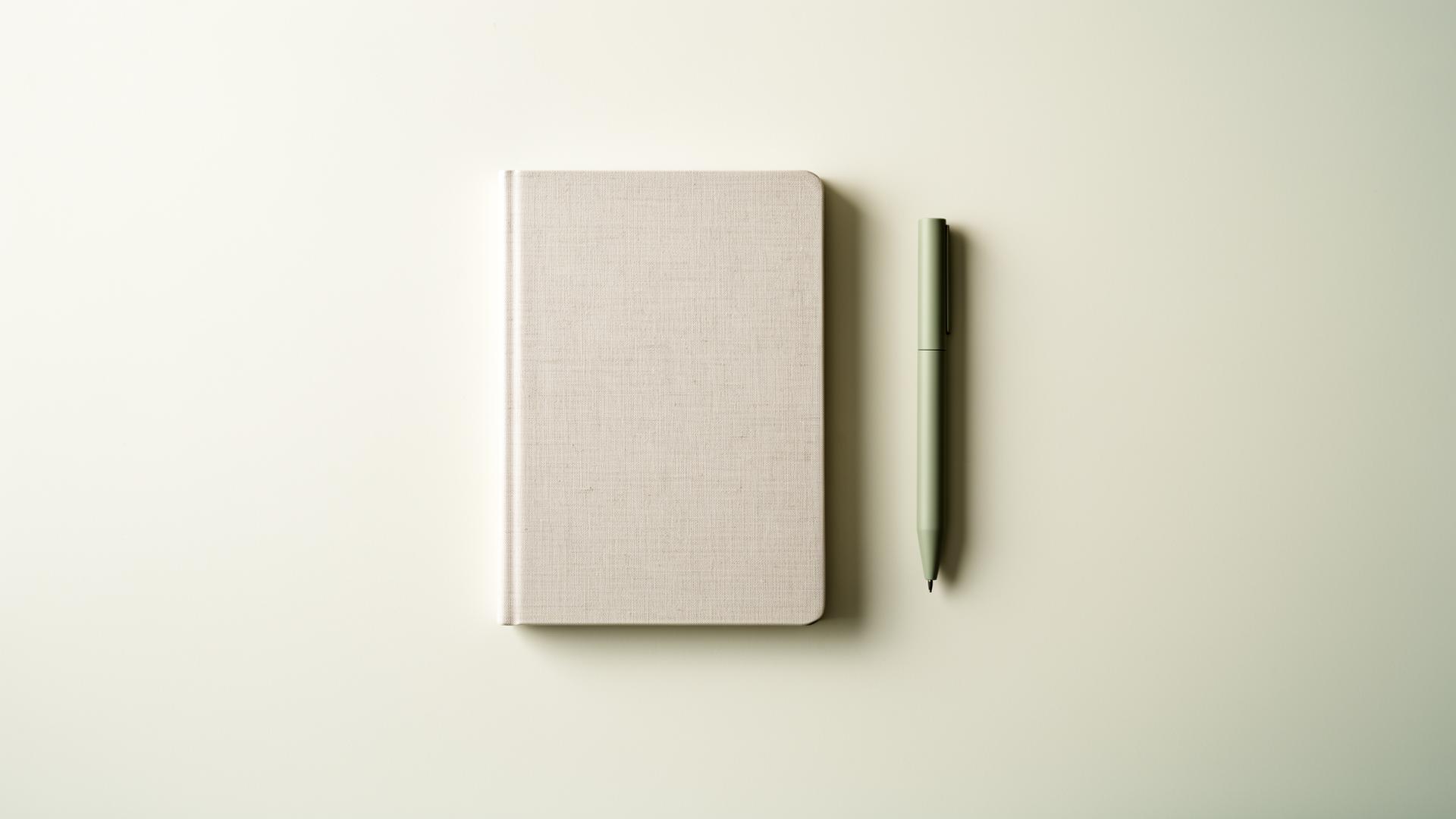 A linen notebook and sage green pen on a clean pale surface — considered, expert work.